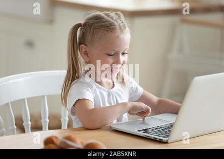 Felice carino piccola ragazza sedersi al tavolo della cucina con laptop Foto Stock