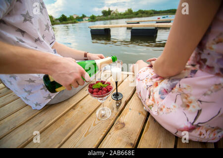 Data romantica sorpresa. Un ragazzo e una ragazza su un molo in legno. Il ragazzo versa dello Champagne nei bicchieri Foto Stock