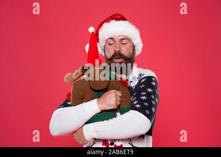 Natale concetto prep. Tanga faccia barbuta inverno maglione. Vacanze invernali. Giocattolo morbido. Abbraccio e l'amore. Acquistare regali. Inverno vendita. Festa d'inverno. La renna. L'uomo santa hat tenere regalo di Natale sfondo rosso. Foto Stock