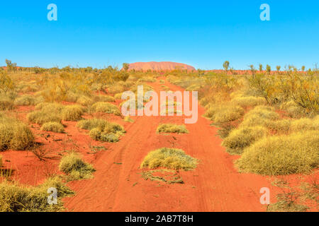 Rosso sentiero di sabbia in secco paesaggio bush in Australian Outback con Ayers Rock in distanza, Uluru-Kata Tjuta National Park, UNESCO, Australia Foto Stock
