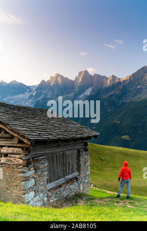 Vista posteriore dell'uomo fuori di una capanna di pietra guardando al Piz Cengalo e Badile, Tombal, Val Bregaglia, Canton Grigioni, Svizzera, Europa Foto Stock