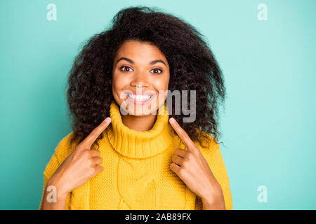 Closeup photo incredibile di pelle scura curly lady indicando le dita sui denti perfetti consigliando dentista giallo indossare pullover a maglia blu isolato teal Foto Stock