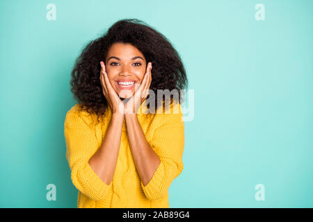 Closeup photo incredibile di pelle scura lady tenendo le mani sul zigomi contentissimo usura giallo pullover a maglia blu isolato teal colore di sfondo Foto Stock