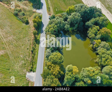 Vista aerea del grigio di una stretta asfaltata country road accanto ad un laghetto con acqua verde circondato da alberi. Foto Stock