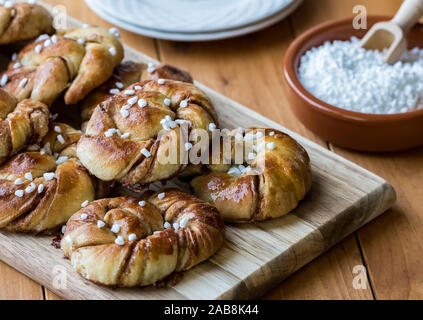 Una vista dall'alto in basso di un pannello di legno con un nuovo lotto di Swedish ciambelle alla cannella (kanelbullar) con un piccolo piatto di perla di zucchero. Foto Stock