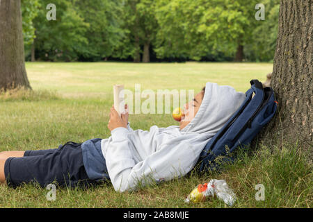 Ragazzo giovane legge un libro e si mangia un Apple presso il parco appoggiata contro la struttura ad albero Foto Stock