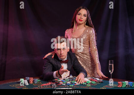 L'uomo gioca a poker al casinò di seduta a tavola con pile di chips, denaro, champagne, carte. Celebrare la vittoria con la donna. Sfondo nero. Close-up. Foto Stock