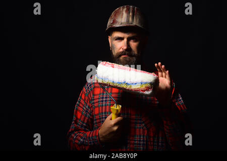 Il lavoro e la riparazione di concetto. Lavoratore con brutale immagine sporca indossa il casco e mantiene il rullo di vernice. Builder o pittore con folta barba. Uomo con volto sorridente espressione isolato su sfondo nero Foto Stock