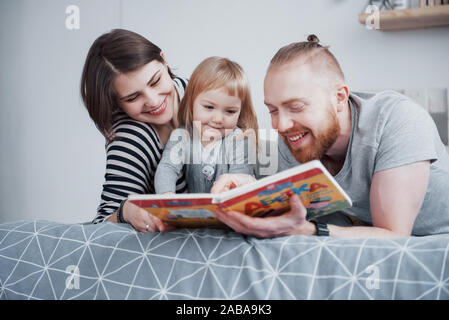 Padre, Madre e figlia piccola lettura libro per bambini su un divano nel soggiorno. Felice la grande famiglia di leggere un libro interessante su un giorno di festa Foto Stock