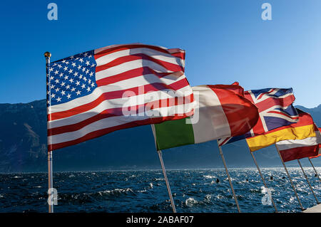 Gruppo di bandiere internazionali sventolare nel vento con il lago di Garda e del monte Baldo sullo sfondo. Limone sul Garda Village, Lombardia, Veneto Foto Stock