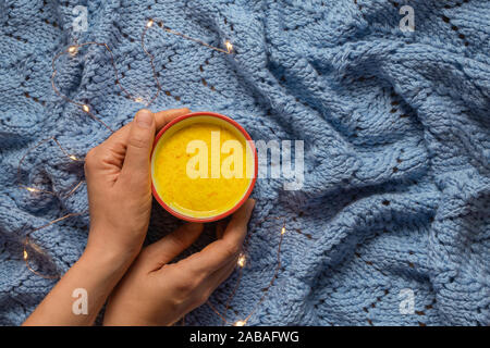 Donna mani tenendo una tazza di wam casalinga curcuma golden latte sulla maglia blu con uno sfondo con le luci fairy. Foto Stock