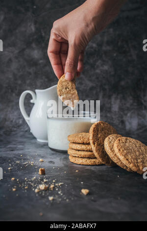 Donna di immersione in mano un cookie in un bicchiere di latte. La prima colazione dolce concetto su un sfondo marmo. I cookie in uno stack con pasticcini sulla tavola di marmo. Foto Stock