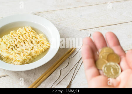 Tagliatelle istante in bianco ciotola, bacchette e mano che trattiene le monete in un bianco tavolo in legno. Concetti di povertà, malnutrizione e cibo a buon mercato. Foto Stock