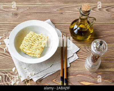 Non cotti tagliatelle istante nella ciotola bianco prima di versare acqua bollente. Olio d'oliva, pepe mill e bastoncini su di un tavolo di legno. La convenienza dei fast food. Foto Stock