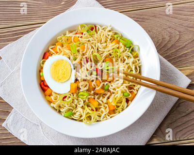 Close-up di spaghetti istantanei con uova e verdure in bianco ciotola e bacchette su un marrone tavolo in legno. La convenienza dei fast food. Vista dall'alto. Foto Stock