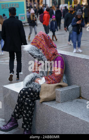 Donna sconosciuta sedette in un quartiere dello shopping con un colorato cappotto a motivi geometrici con il cappuccio che copre la sua faccia, Leeds, West Yorkshire, Inghilterra, Regno Unito. Foto Stock