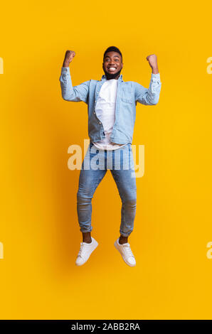 Euforico ragazzo afro salti in aria, celebrando il successo su sfondo giallo Foto Stock