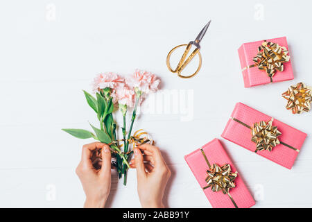 Piano di composizione laici mani femminili rendendo bouquet accanto a confezioni regalo e un paio di forbici su sfondo bianco, vista dall'alto. Foto Stock