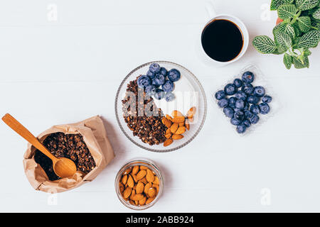 Ciotola di muesli al cioccolato con mirtilli, mandorle e yogurt accanto alla tazza di caffè sul tavolo bianco, vista dall'alto. Foto Stock