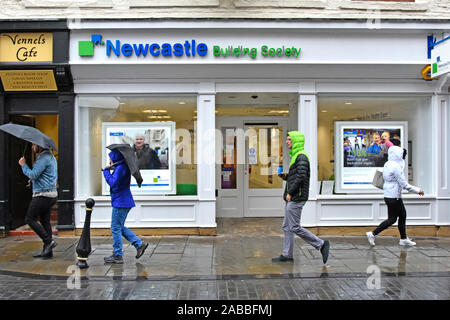 Scena di strada & studio candidi persone vestite per affrontare una giornata di pioggia camminando lungo il marciapiede bagnato alcuni ombrelloni nella strada dello shopping di Durham Regno Unito Inghilterra Foto Stock