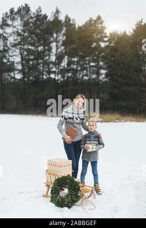 La famiglia felice, la madre e il suo piccolo figlio, con un regalo di Natale scatola in foresta d'inverno. Elegante in legno con slitta presenta e ghirlanda di Natale sul Foto Stock