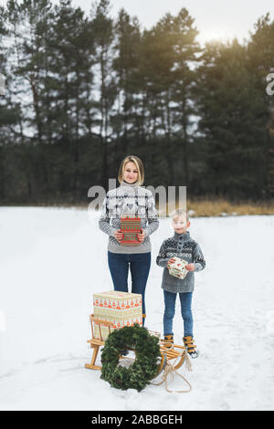 La famiglia felice, la madre e il suo piccolo figlio, con un regalo di Natale scatola in foresta d'inverno. Elegante in legno con slitta presenta e ghirlanda di Natale sul Foto Stock