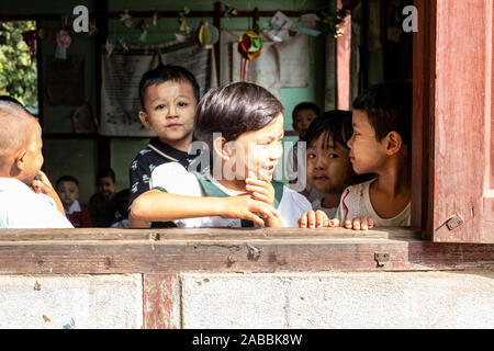 Gli studenti della scuola elementare lean al di fuori della loro classe finestra con sorrisi e curiosità in Kanne villaggio sul fiume Chindwin del Myanmar (Birmania) Foto Stock