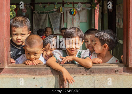 Gli studenti della scuola elementare lean al di fuori della loro classe finestra con sorrisi e curiosità in Kanne villaggio sul fiume Chindwin del Myanmar (Birmania) Foto Stock