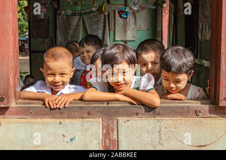 Gli studenti della scuola elementare lean al di fuori della loro classe finestra con sorrisi e curiosità in Kanne villaggio sul fiume Chindwin del Myanmar (Birmania) Foto Stock