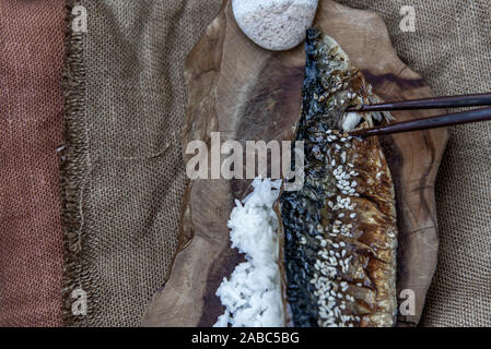 La mano è utilizzando bastoncini per prelevare la saba grigliate di pesce sul riso con salsa teriyaki su un sacco di sfondo, stile giapponese. Foto Stock