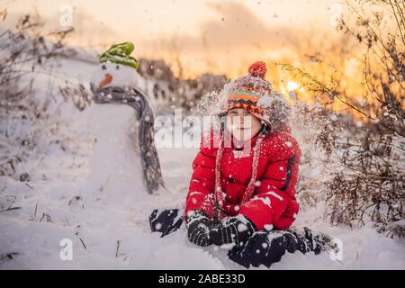 Carino boy in rosso abbigliamento invernale costruisce un pupazzo di neve. Divertimento invernale Outdoor Concept Foto Stock