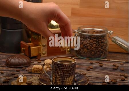 Ragazza di mano mette un pezzo di zucchero di canna in una tazza di caffè, i chicchi di caffè e lo zucchero di canna, macinino da caffè su uno sfondo di legno Foto Stock