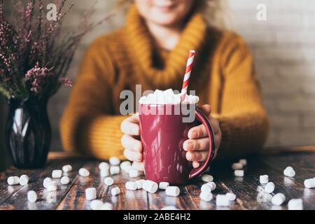 Donna in felpa gialla azienda cioccolata calda tazza coperta con marshmallow sul legno scuro dello sfondo. Foto Stock