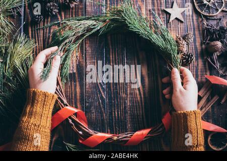 Mani femminili rendendo ghirlanda di Natale con abete brunch scuro su uno sfondo di legno Foto Stock