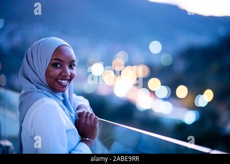Moderna africana donna musulmana in notte al balcone sorridente in telecamera con city bokeh luci in background Foto Stock