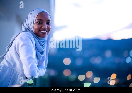 Moderna africana donna musulmana in notte al balcone sorridente in telecamera con city bokeh luci in background Foto Stock