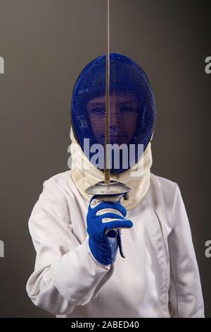 Little Boy fencer armati con una piattina di pinza in bianco le attrezzature sportive e di un casco protettivo su sfondo scuro Foto Stock