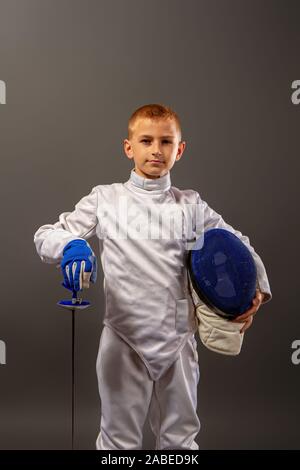 Little Boy fencer armati con una piattina di pinza in bianco le attrezzature sportive e di un casco protettivo su sfondo scuro Foto Stock