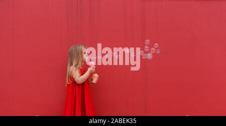 Ragazza bionda bambino in abito rosso su uno sfondo della parete soffia bolle. Foto Stock