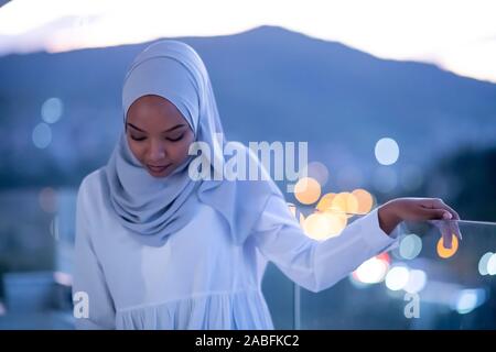 Moderna africana donna musulmana in notte al balcone sorridente in telecamera con city bokeh luci in background Foto Stock