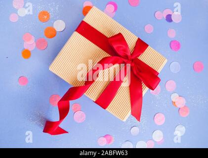 Giftbox legato con red ribbon colore su sfondo viola con sparklets e coriandoli. Piatto stile di laici. Foto Stock