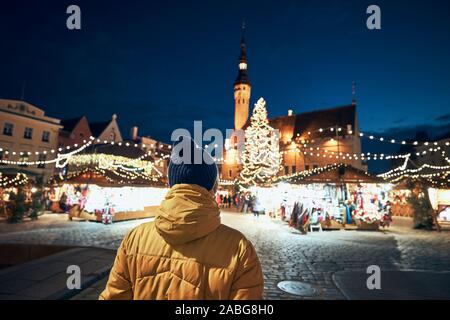 Vista posteriore dell'uomo in un abbigliamento caldo durante la notte a piedi in città. Tradizionale mercatino di Natale in piazza del Municipio di Tallinn, Estonia. Foto Stock