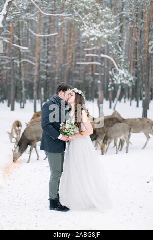 Amare giovane a piedi, abbracciando e baciando, in inverno boschi innevati. Allevamento di cervi sullo sfondo. Inverno di nozze. Foto Stock