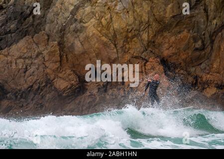 Surfer in wetsuit a cavallo di un onda sulla tavola da surf pericolosamente vicino a rocce di scogliera sul mare A la Pointe du Raz, Finistère Bretagna, Francia Foto Stock