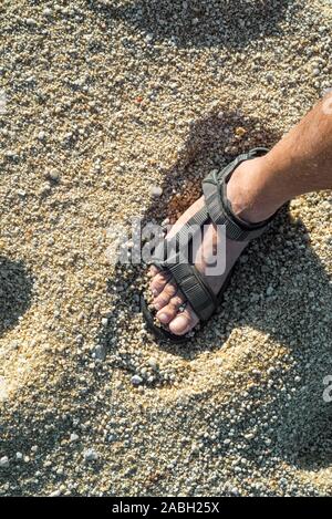 Close-up del piede maschile in sandalo permanente sulla spiaggia con mare grosso granelli di sabbia Foto Stock