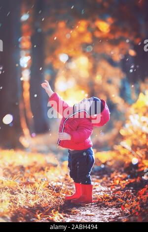 Poco toddler boy in rosso stivali di gomma e giacca rossa la cattura di gocce di pioggia in autunno park. Arancio foglie della foresta sullo sfondo Foto Stock