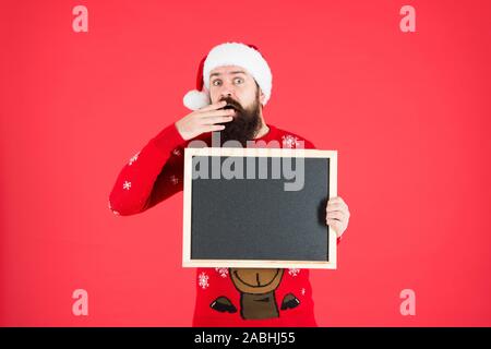 Incredibile. Annuncio d'inverno. Evento invernale. Vacanze invernali. Santa Claus hat con la pubblicità. Lavagna per informazioni. Man tenere lavagna vuota copia spazio. Guy babbo natale sfondo rosso. Foto Stock