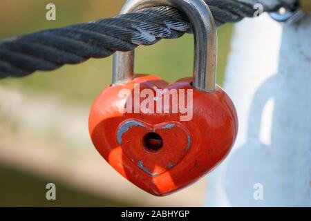 Il dispositivo di bloccaggio rosso appendere su un cavo su un ponte. Simbolo di amore e fedeltà eterna metal heart-a forma di lucchetto sul ponte di close-up. Tradizioni di matrimonio. Foto Stock