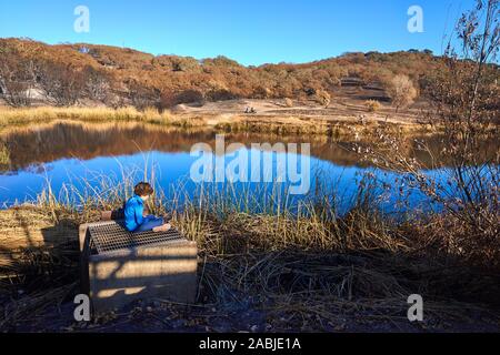 Giovane ragazzo in blu giacca e pantalone si siede vicino a un lago. Bruciò ance, erba e alberi a Foothill Parco Regionale in Windsor, Sonoma County, California. Foto Stock