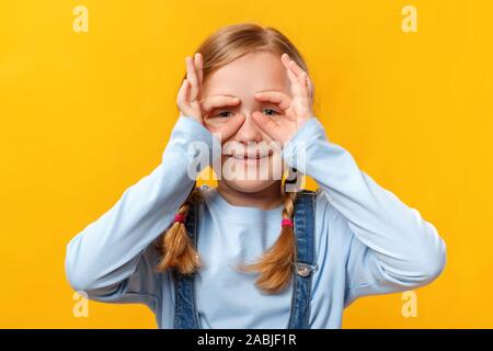 Funny bambino è divertente e rende i bicchieri di dita. Bambina in una maglietta blu su sfondo giallo. Foto Stock